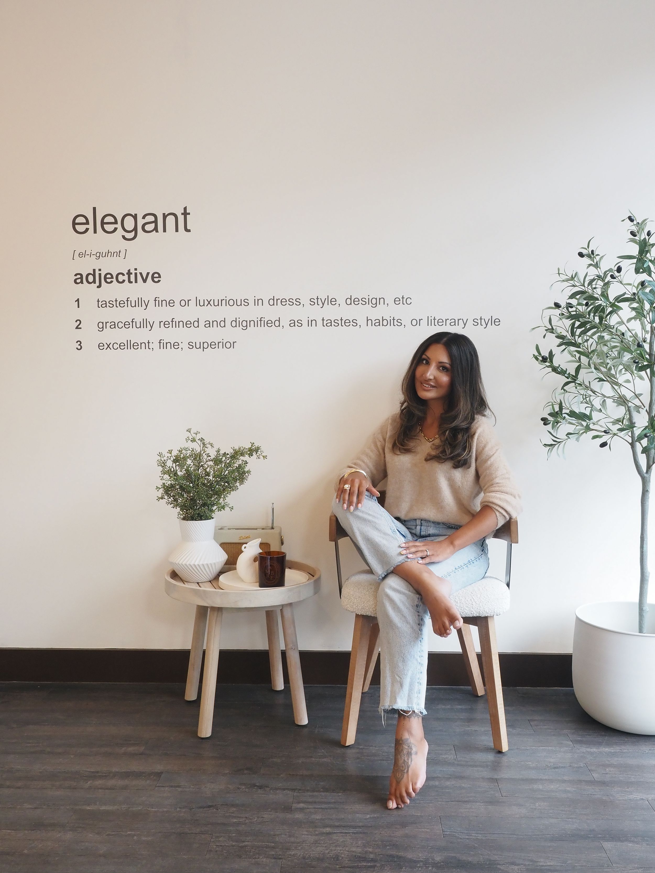 Modern minimalist room setting with someone seated in casual attire by wooden side table with plants and elegant text on wall.