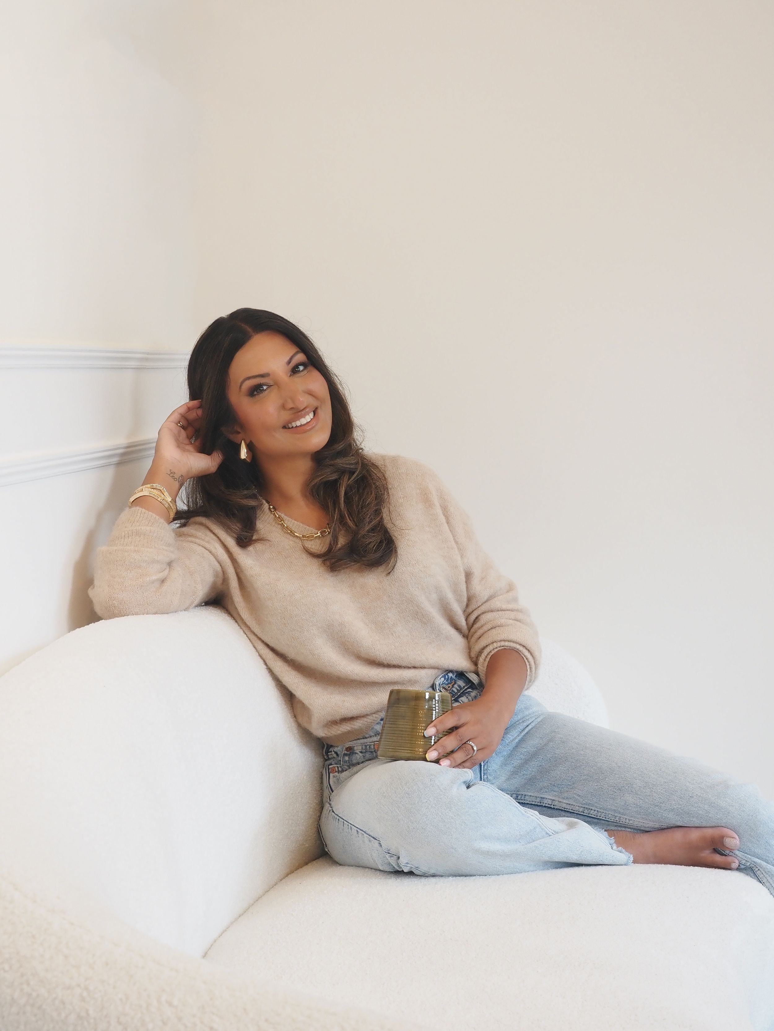 A person relaxes on a white couch wearing a beige sweater and light jeans while holding a drink.