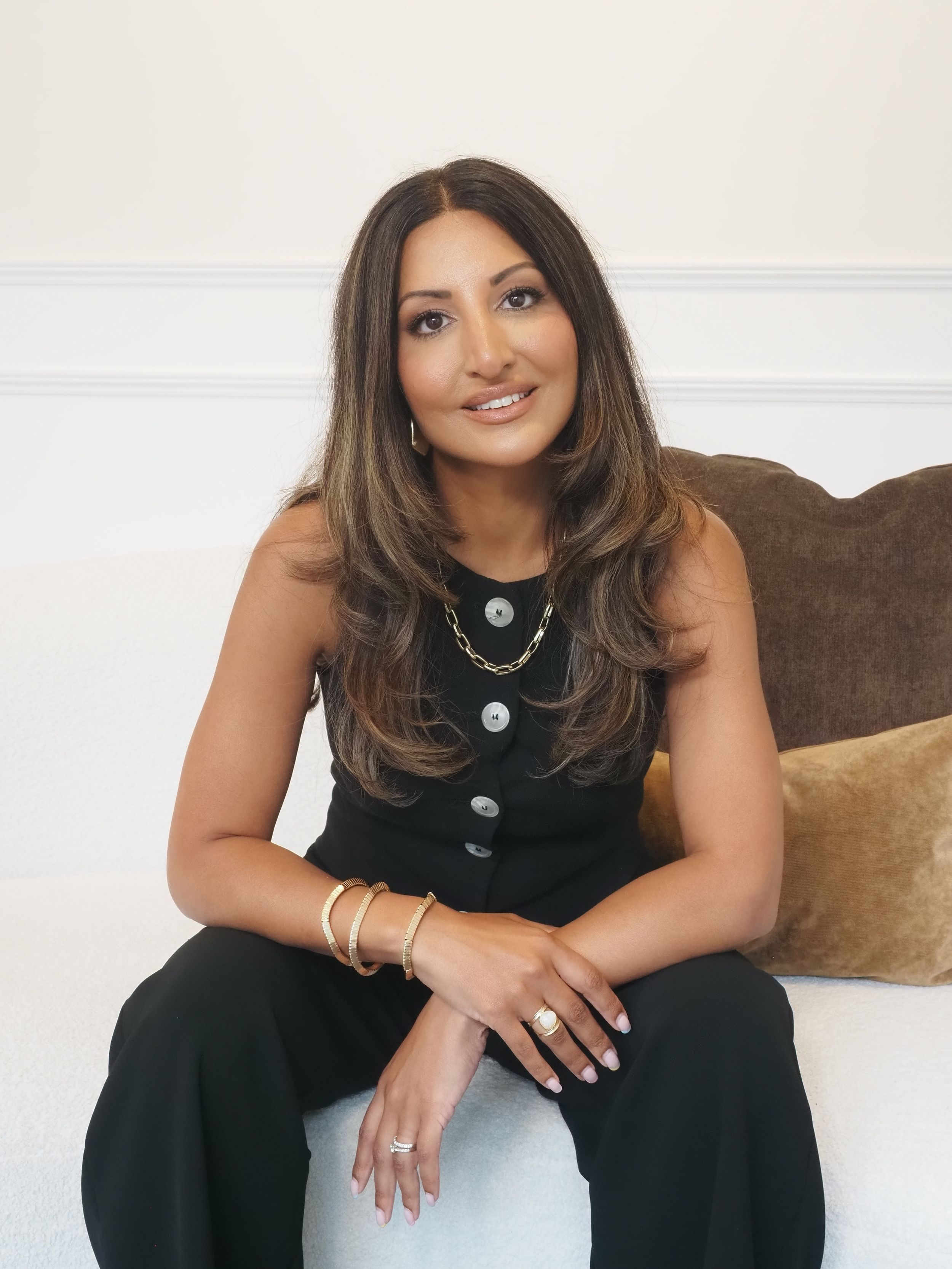 A person in a black top sits elegantly on a couch with bracelets and layered necklaces against a white wall.