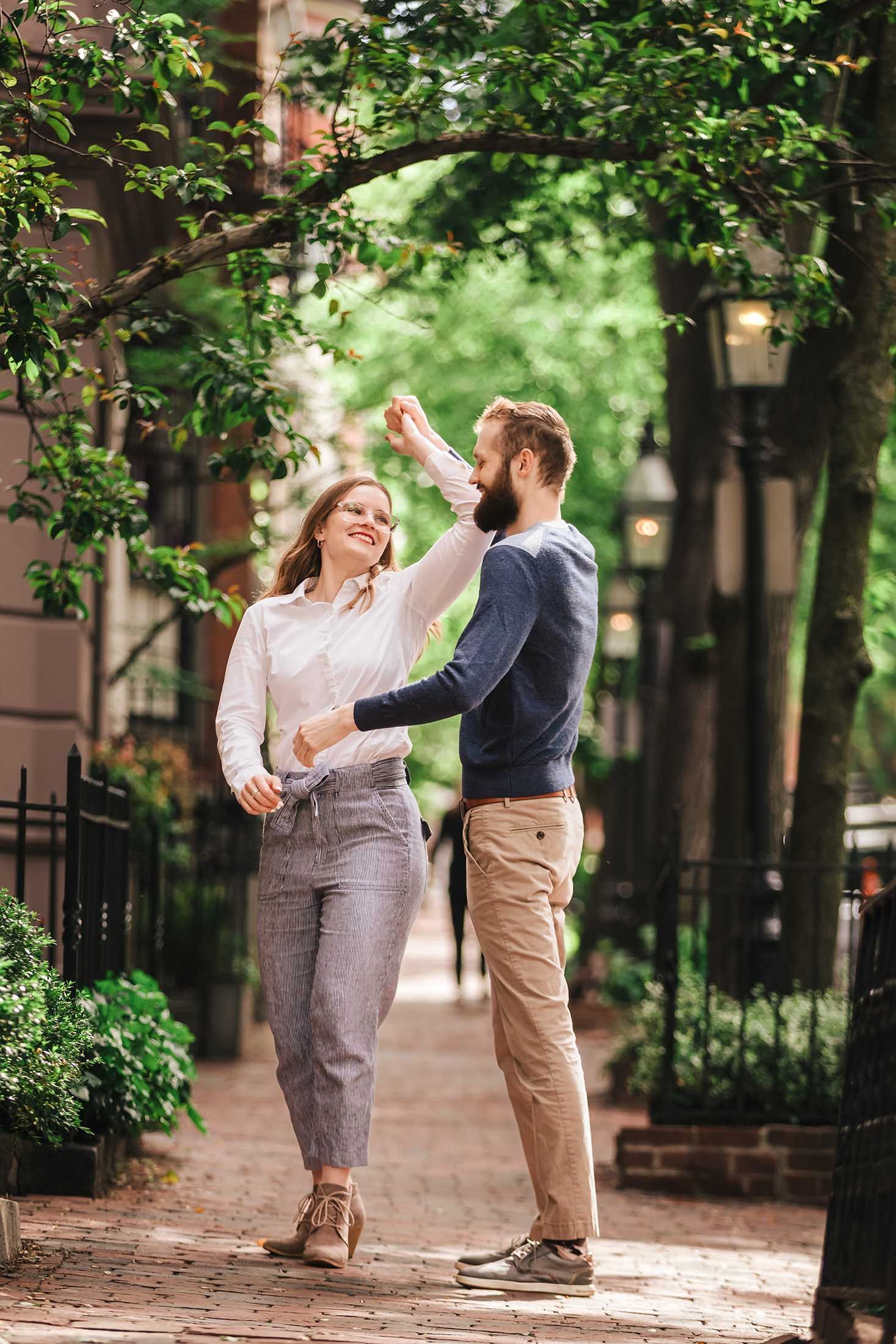 A couple dances playfully on a cobblestone sidewalk in Beacon Hill, surrounded by brick buildings, lush foliage, and vintage street lamps