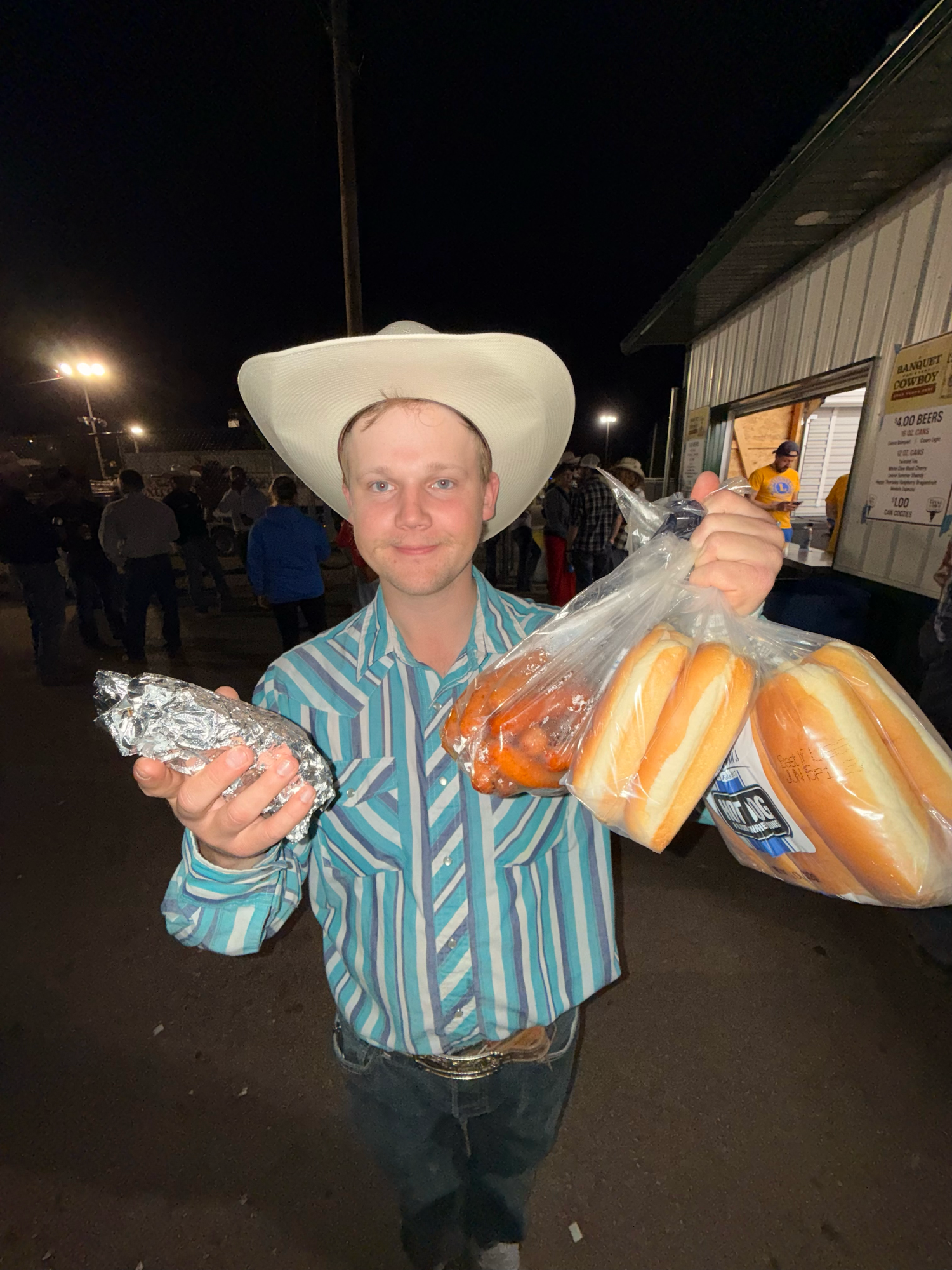 Someone holding food items while wearing a cowboy hat and western shirt at night.