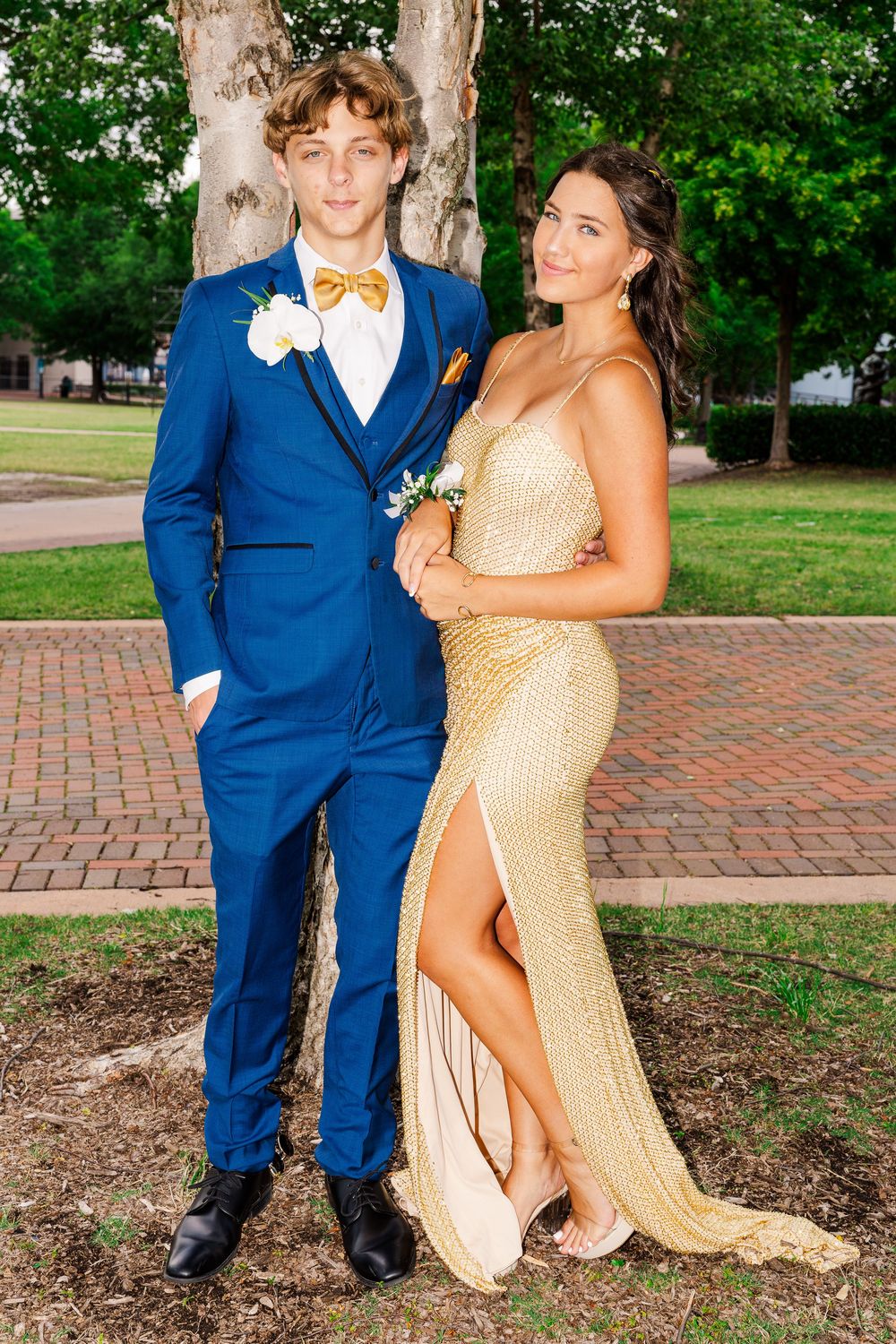 Couple posing in prom attire, blue suit and gold sequin dress outdoors.