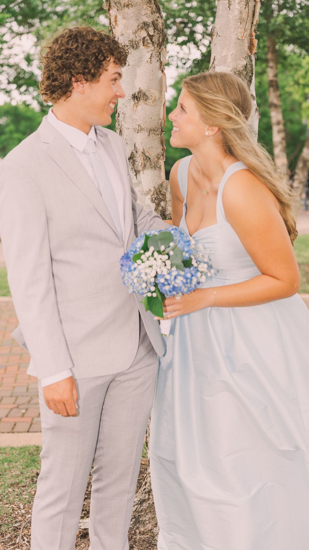 Couple in formal wear smiling at each other holding blue hydrangea bouquet.