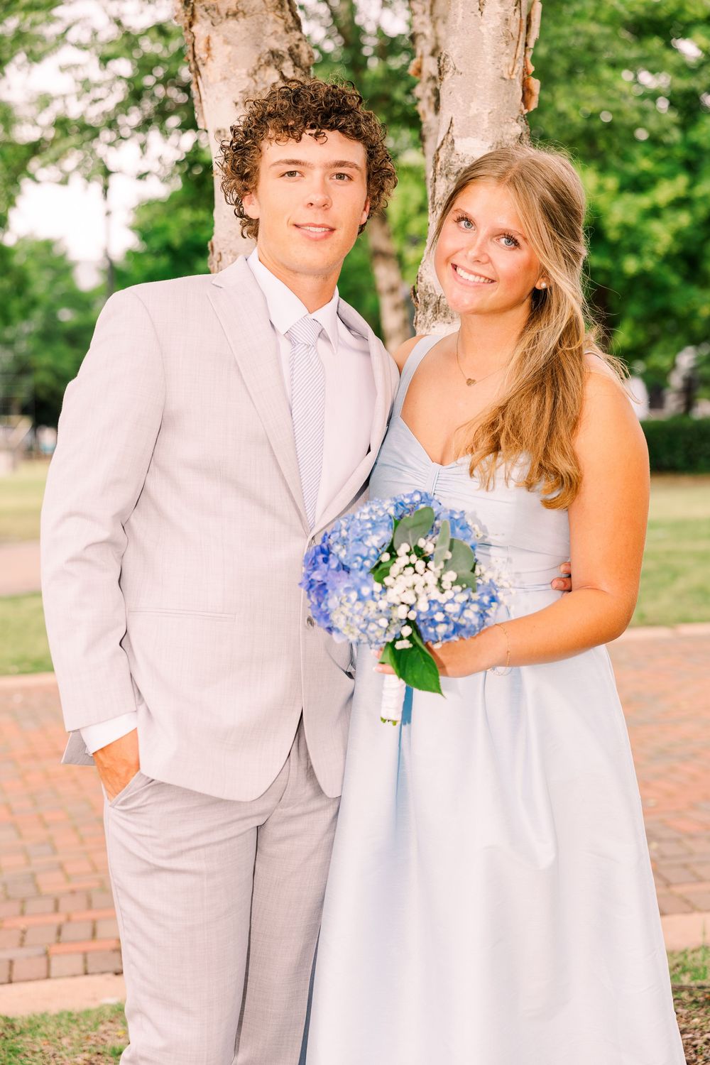 Young couple in formal prom attire with blue hydrangea bouquet outdoors.