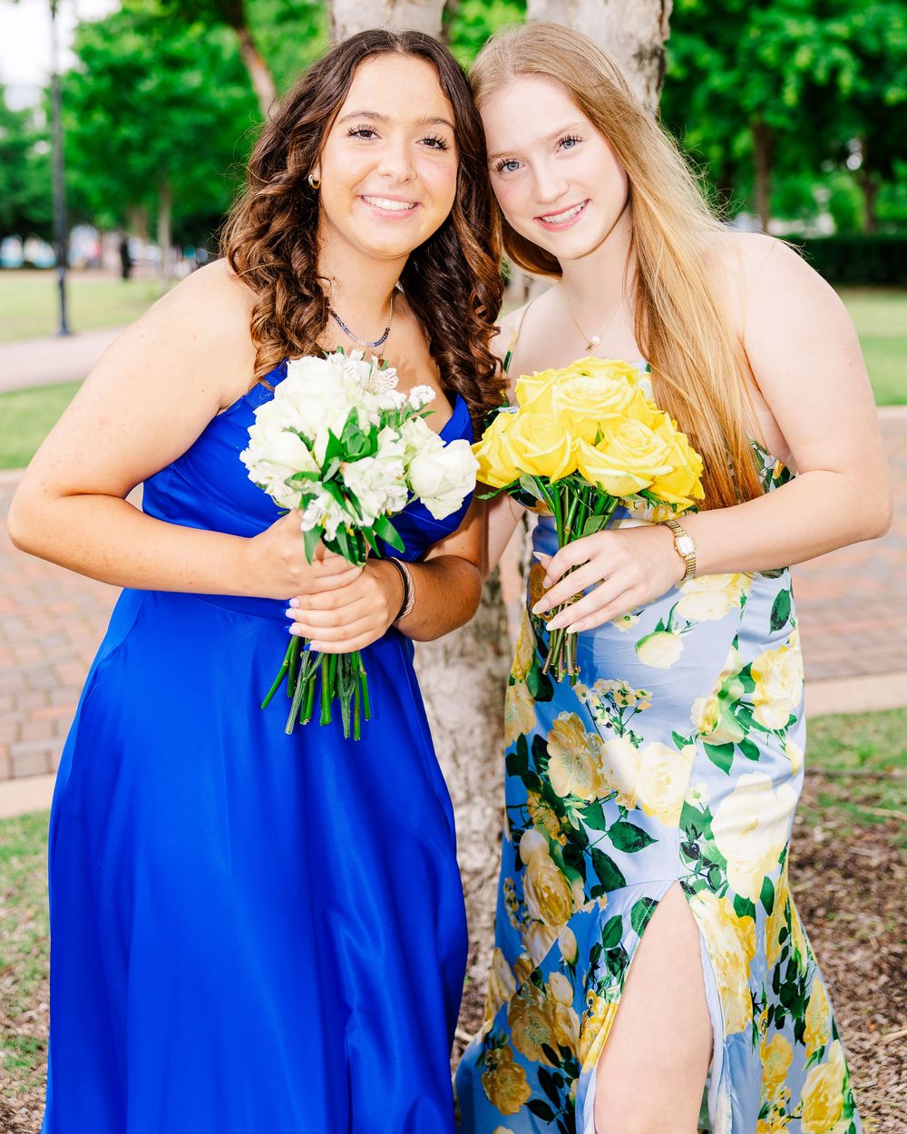 Two girls in prom dresses holding white and yellow rose bouquets outdoors.