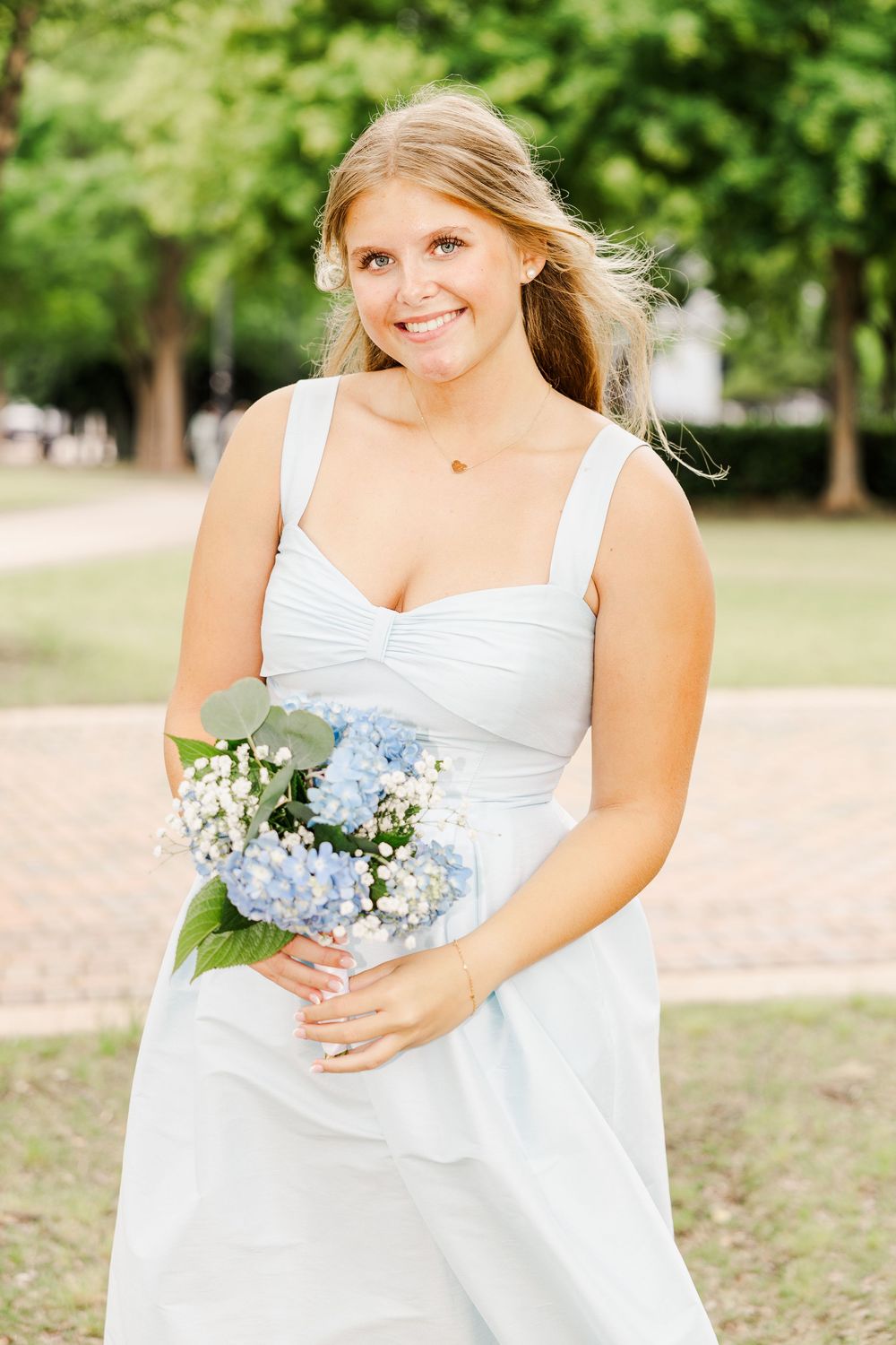 Smiling blonde woman in white dress holding blue hydrangea bouquet outdoors.