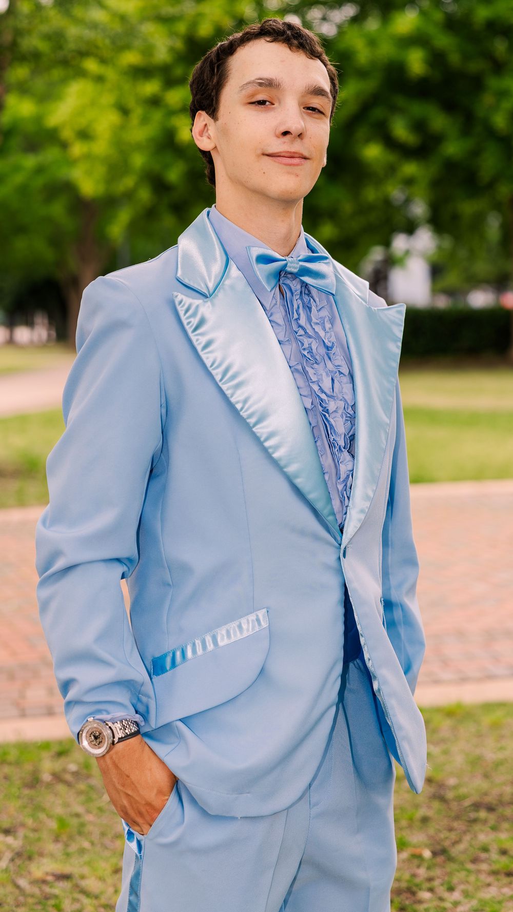 Young man posing in light blue satin tuxedo with bow tie outdoors.