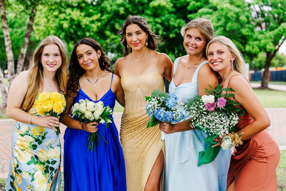 Five young women in colorful formal prom dresses holding bouquets posing outdoors in park.