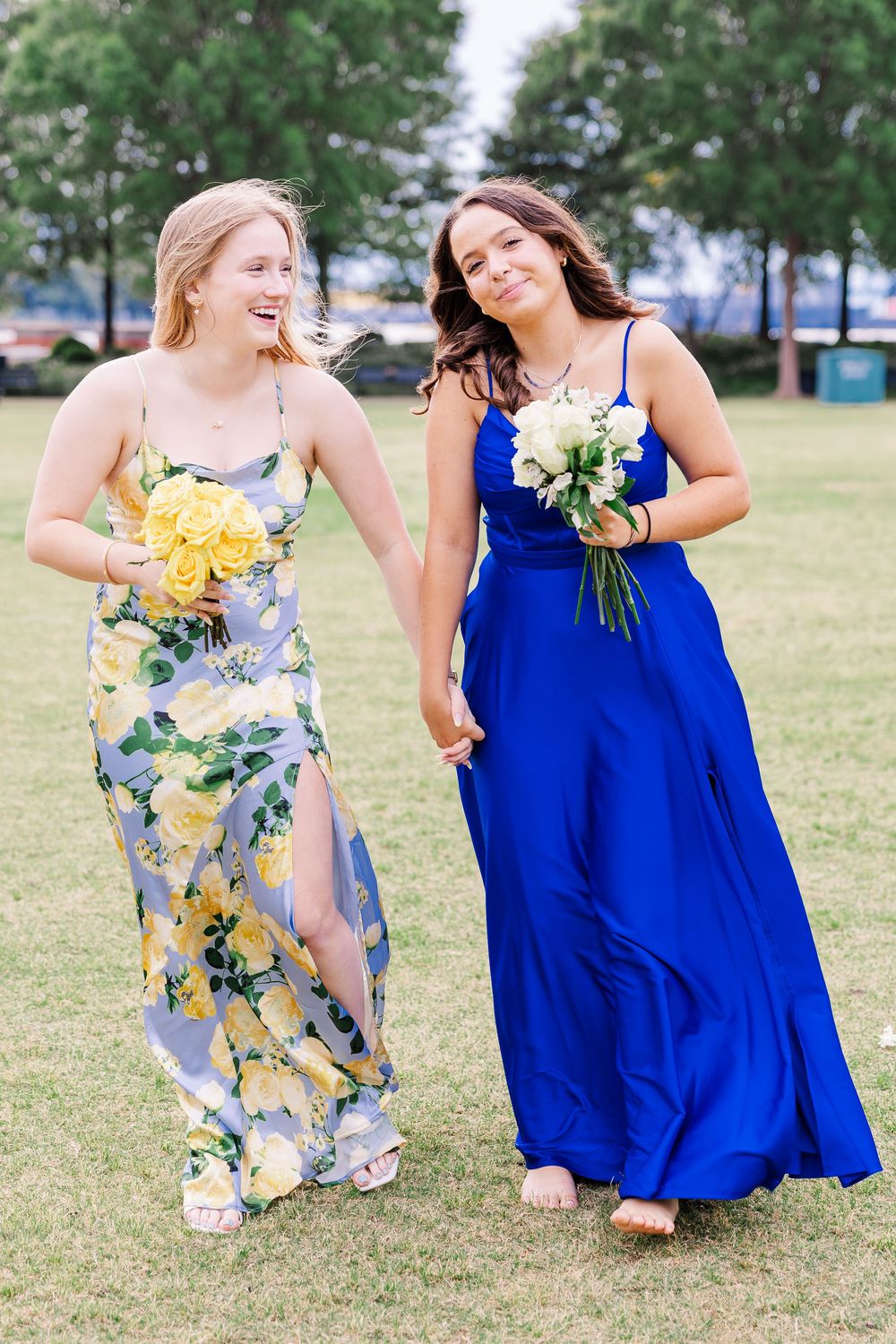 Two smiling girls in floral and blue prom dresses holding flowers walking on grass.