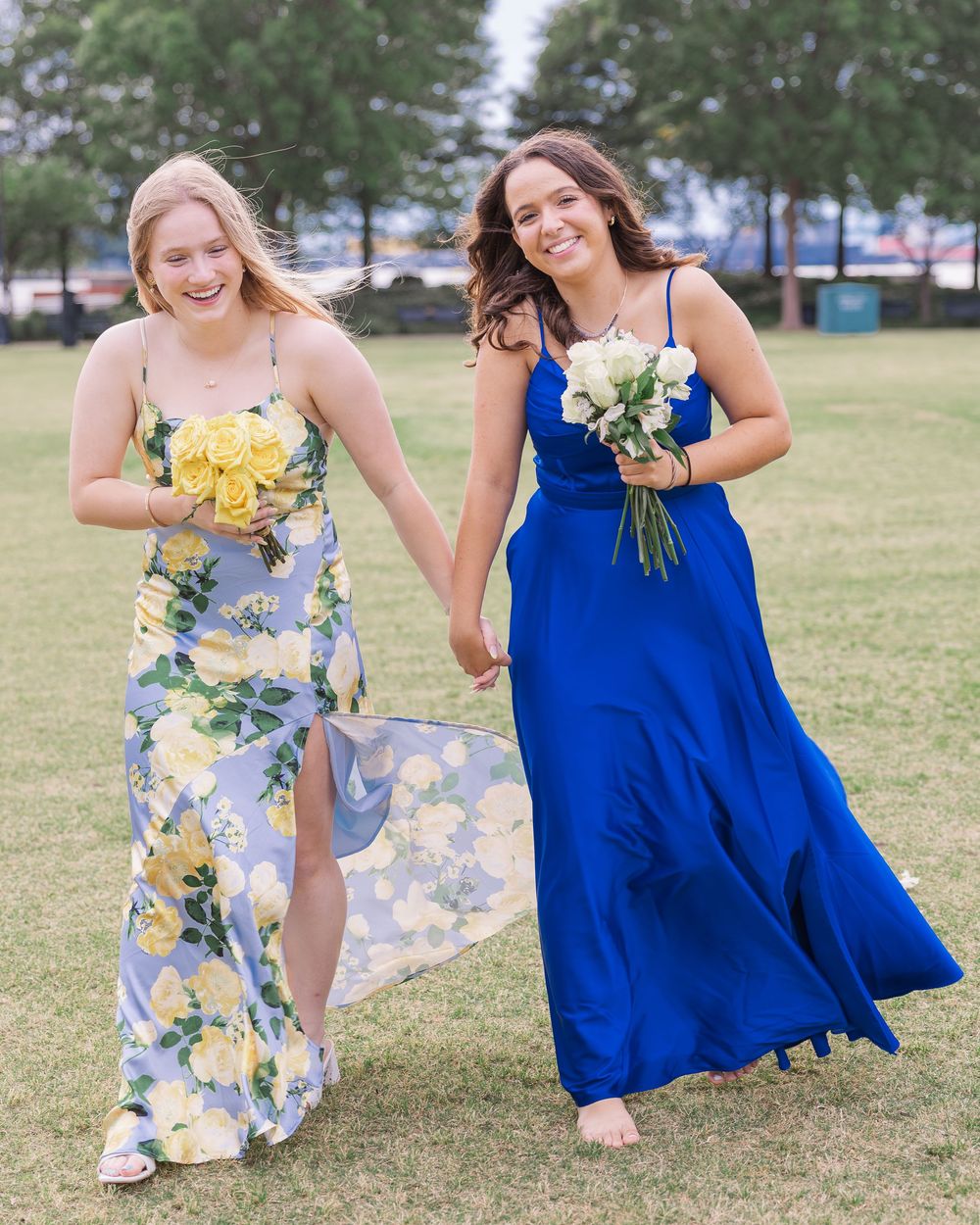 Two smiling girls in floral and royal blue formal dresses holding bouquets in park.