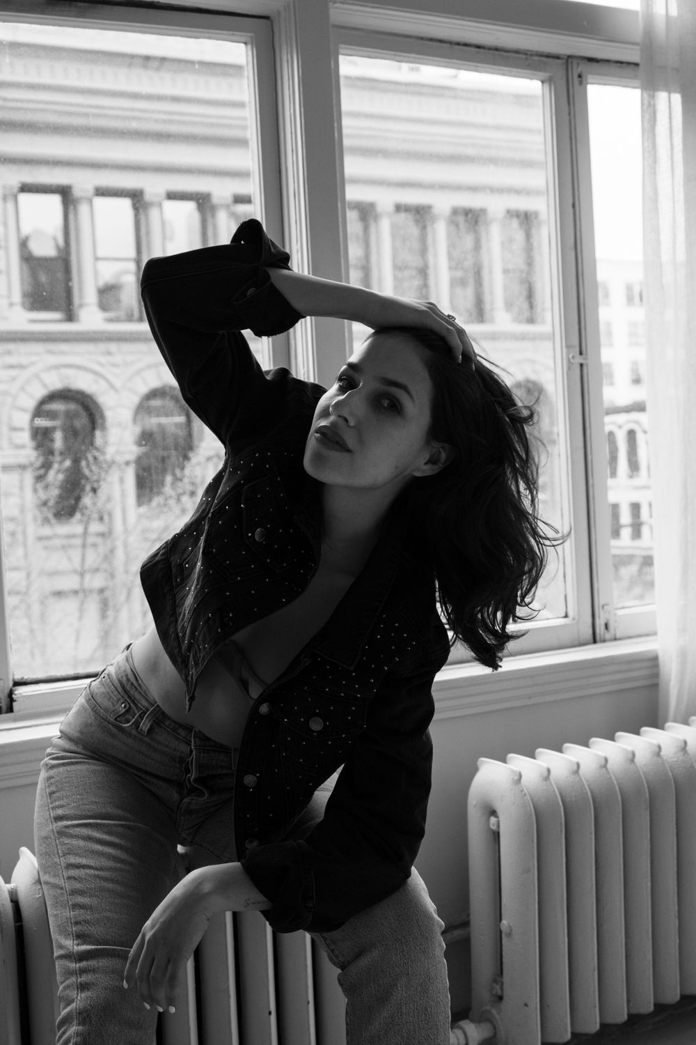 Black and white artistic photograph showing person leaning back by windows with radiators in an empty interior space.