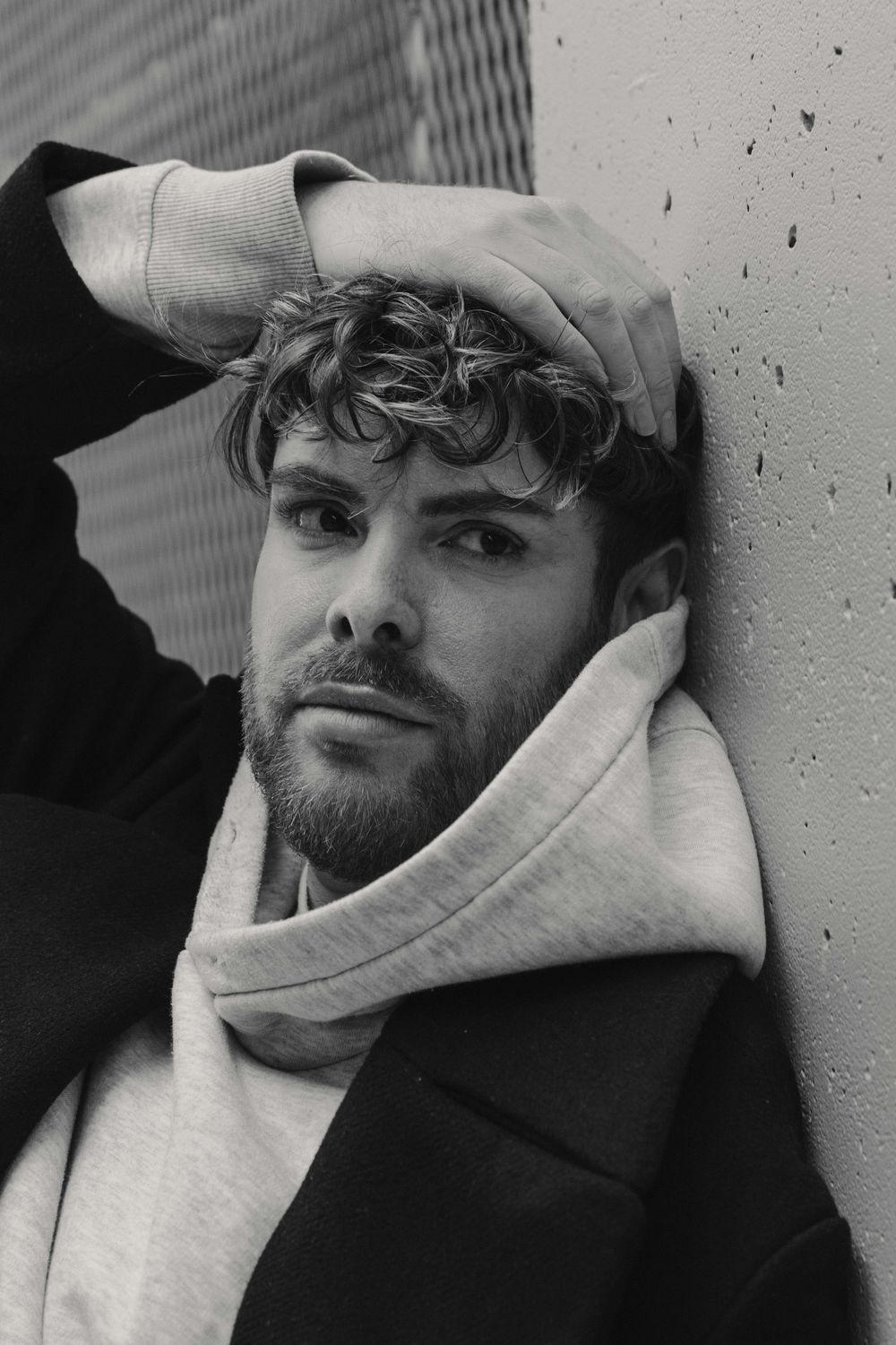 Black and white portrait of someone in a light hoodie and dark coat leaning against a wall.