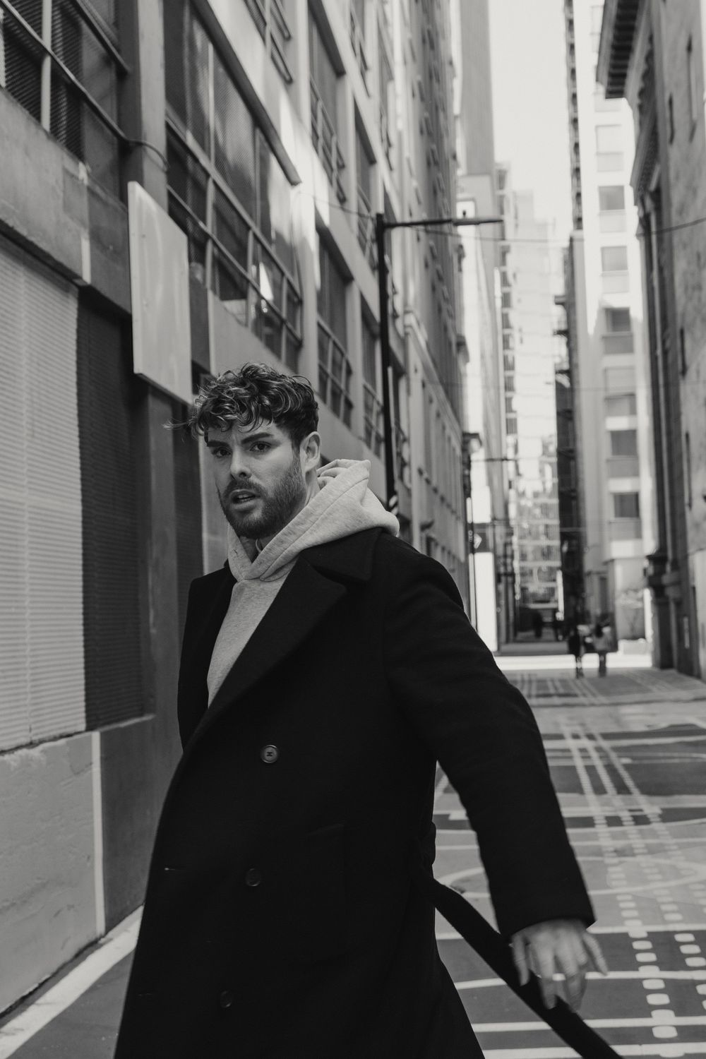 Black and white photo of person in winter coat and hoodie walking in urban alleyway between tall buildings.