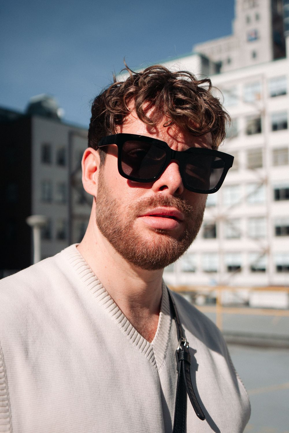 Person wearing white knitted sweater and black sunglasses poses outside urban building on sunny day.