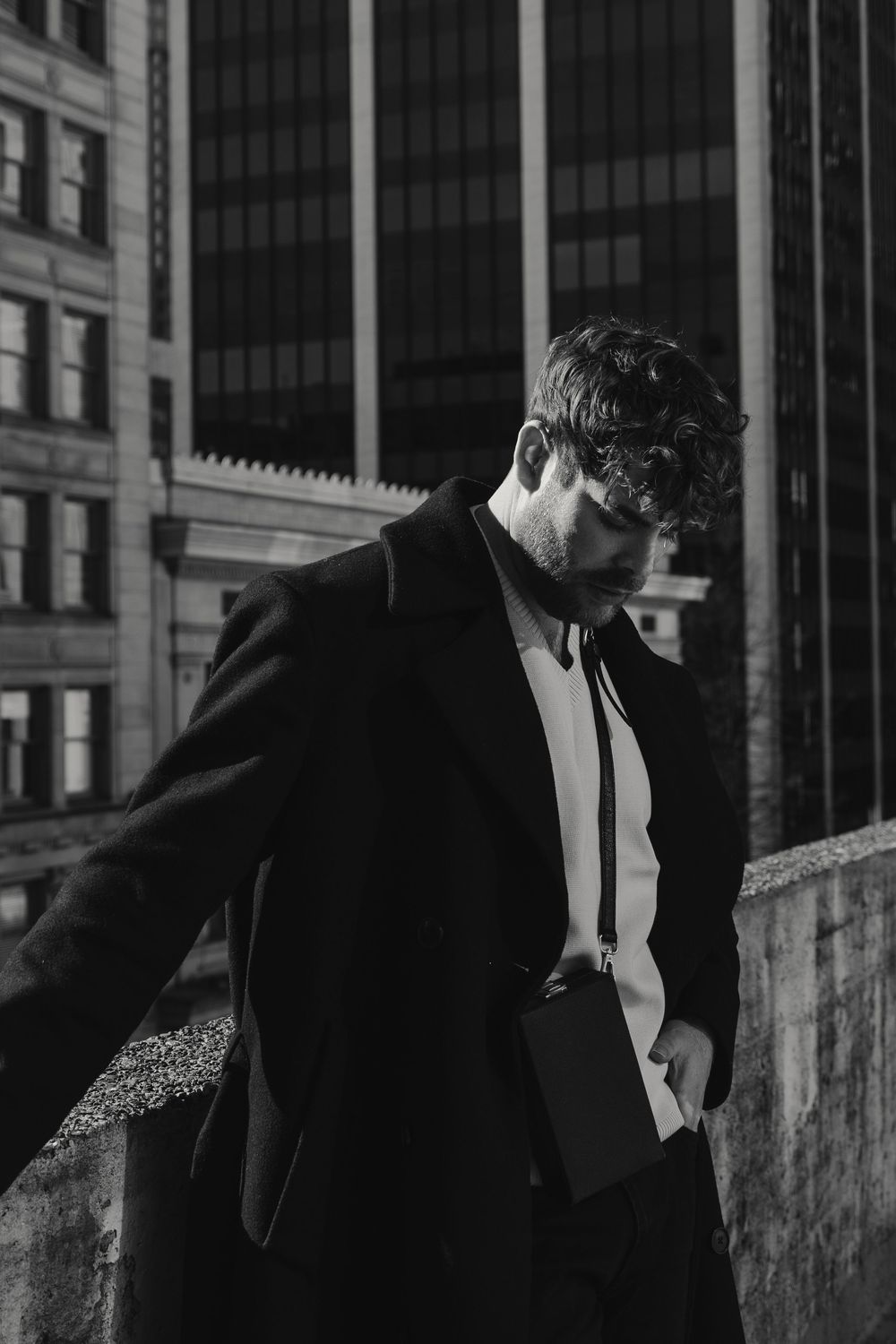 Black and white portrait of someone in a suit and tie looking down against an urban backdrop of office buildings.