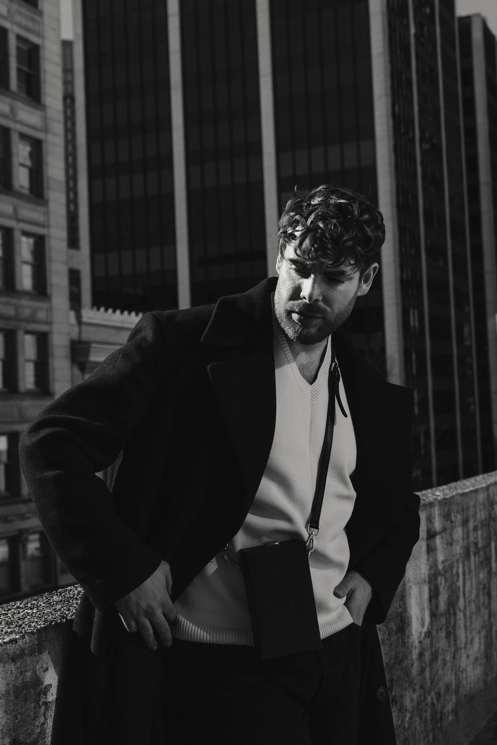 Black and white urban fashion portrait of a person wearing a blazer and cap against a city backdrop with modern buildings.