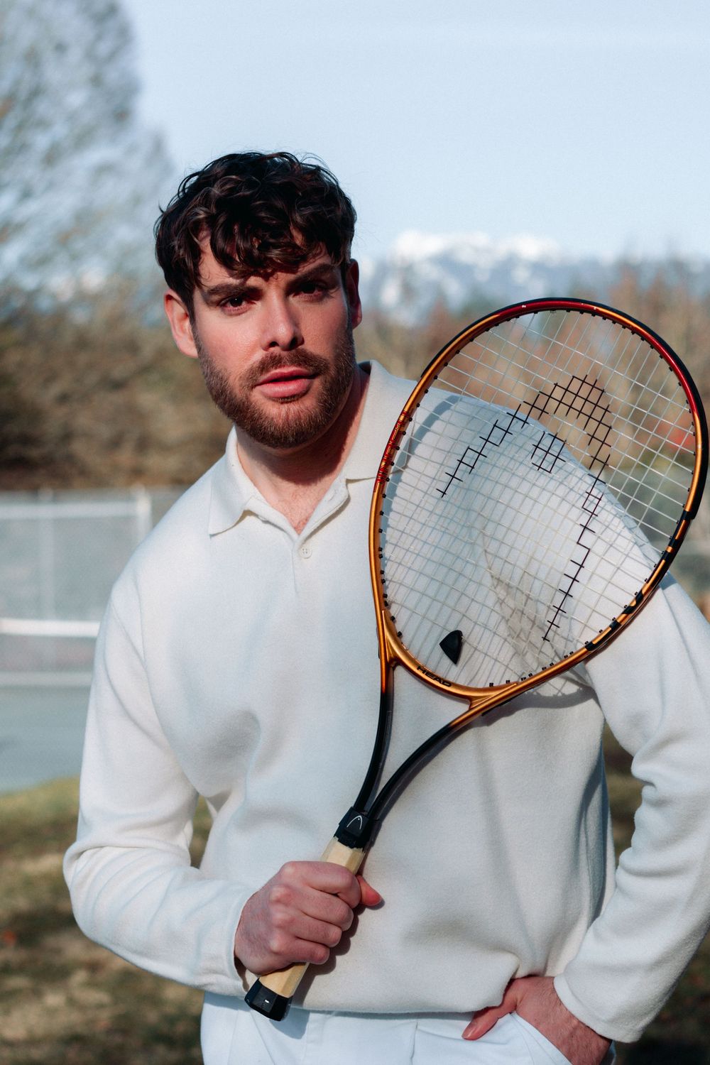 Tennis player in white attire poses with racket on outdoor court with mountains visible in background.