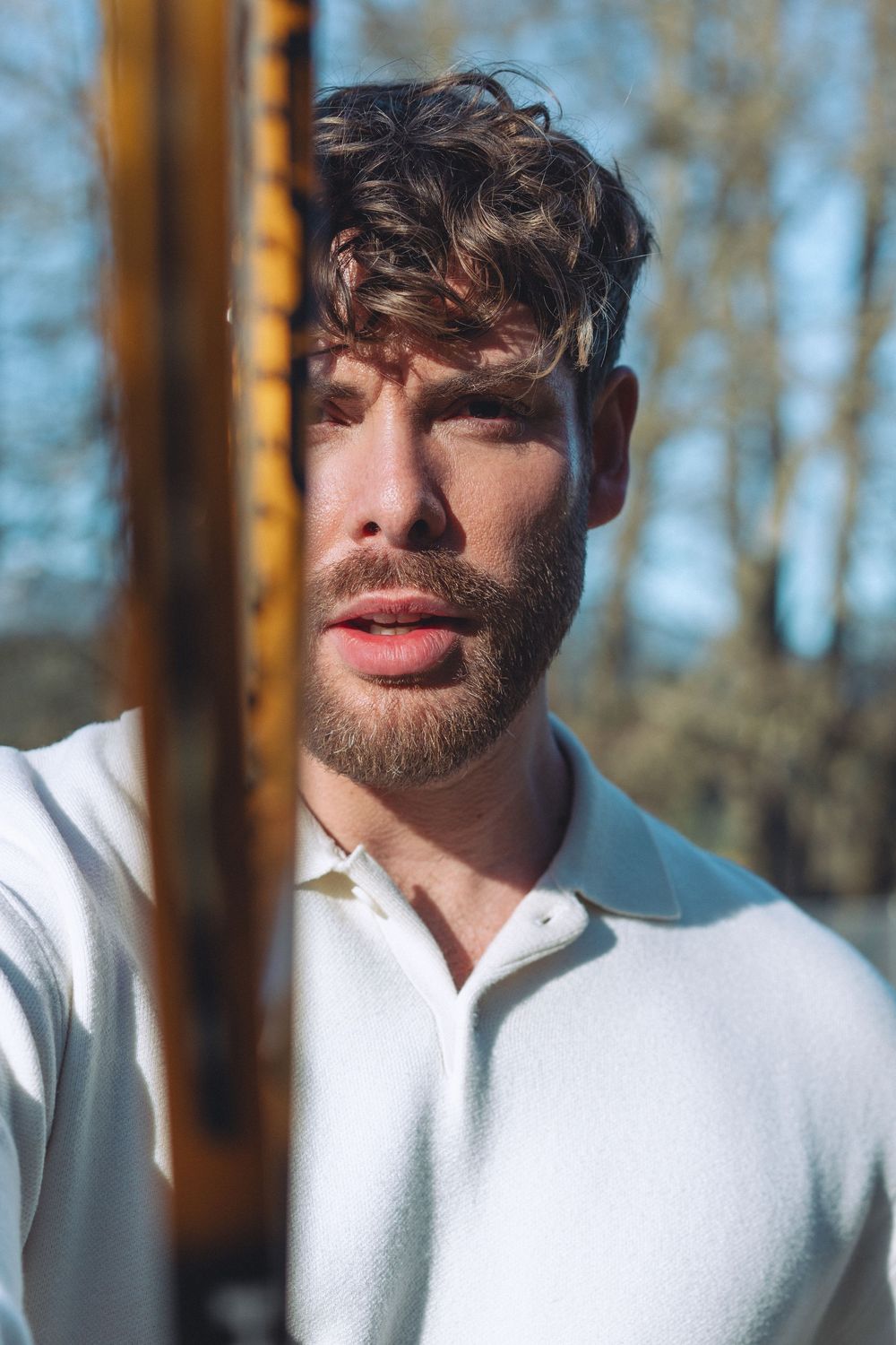 Portrait of a person in a white polo shirt outdoors in natural sunlight with trees in the background.