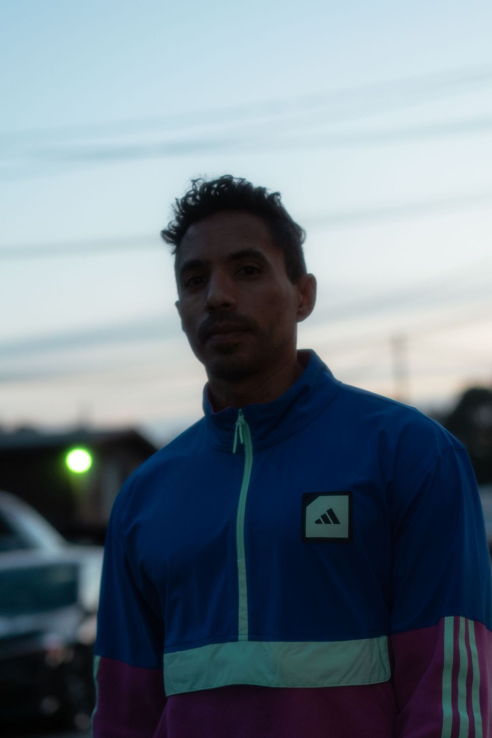 A person wearing a blue and turquoise Adidas track jacket stands outdoors at dusk.