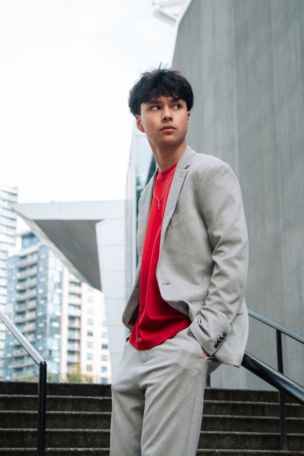 Fashion portrait of someone in a light gray suit and red sweater standing on concrete stairs in an urban setting.