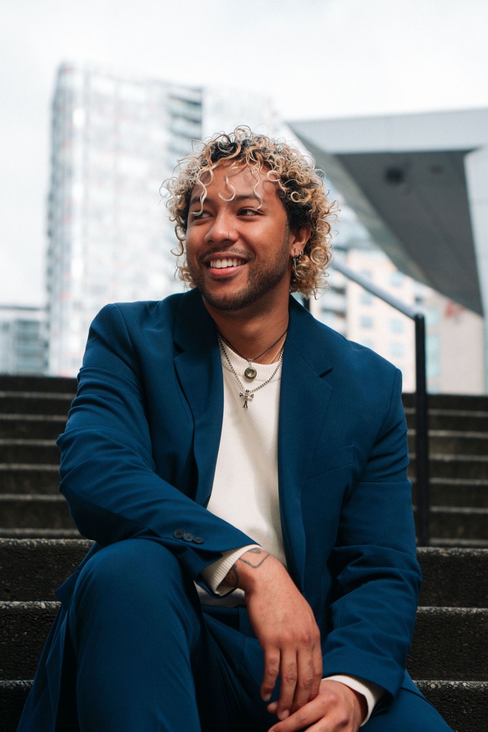 A person with curly hair wearing a navy blue suit and white shirt sits smiling on concrete stairs outdoors.