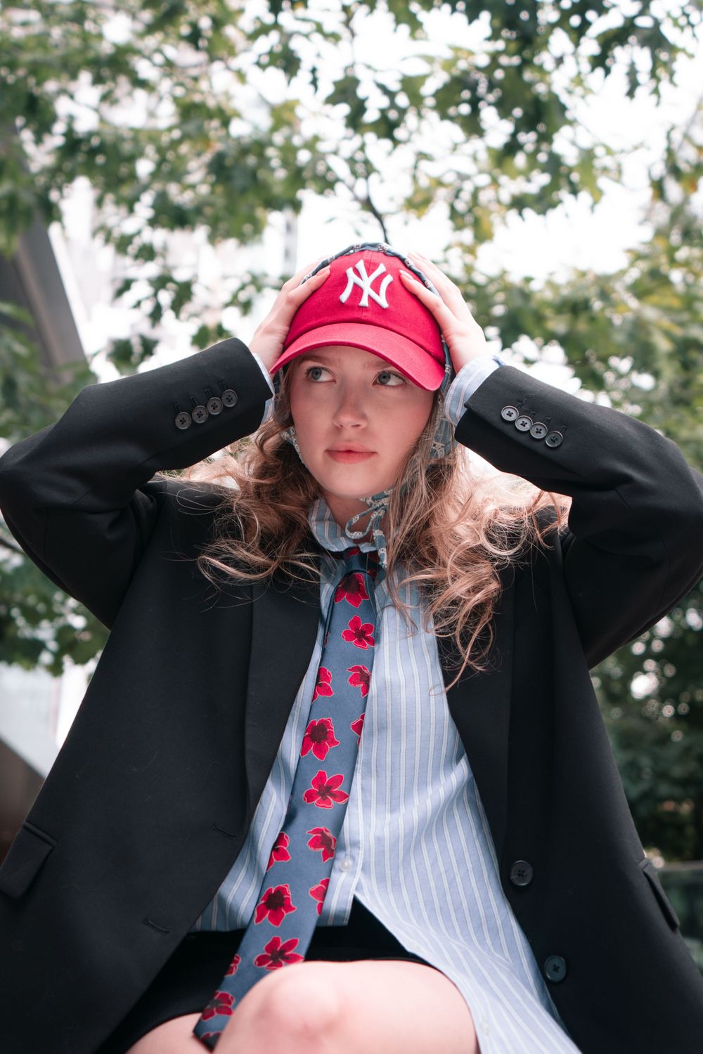 Fashionable outfit featuring red NY Yankees cap, black blazer, striped shirt and patterned tie in outdoor setting with trees.