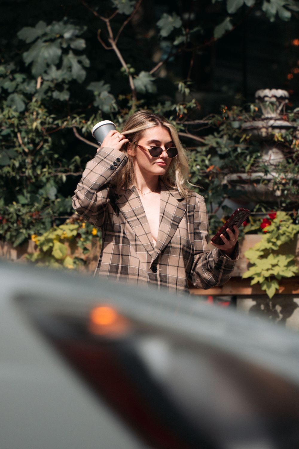 Woman in plaid blazer and sunglasses checks phone while standing outdoors in garden setting.
