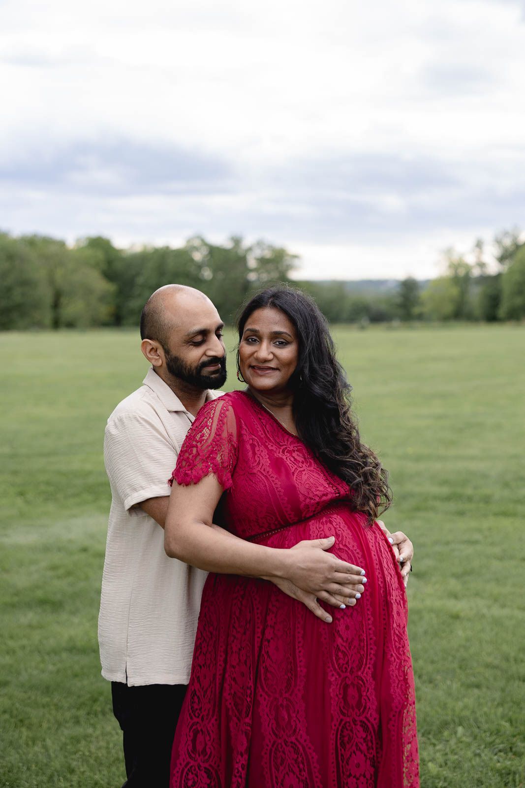 A couple embraces in a green field as they pose for a maternity photo, with the woman wearing a red lace dress.