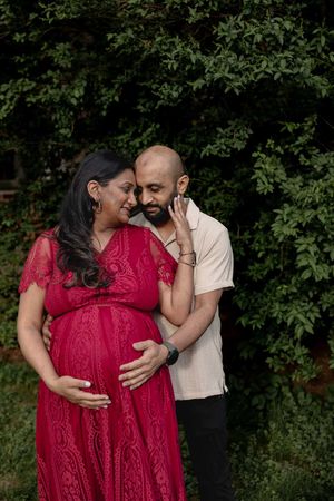 Couple embraces in front of greenery with a woman in red lace dress cradling her pregnant belly.