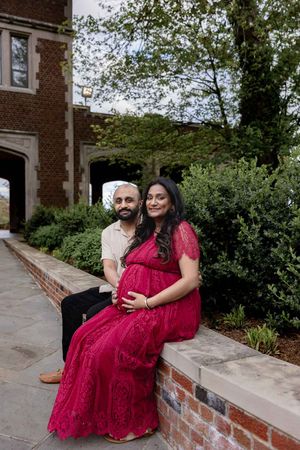 Couple sits on brick ledge by building with woman in red maternity dress holding her baby bump.