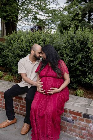 Expectant couple sits on brick ledge in garden as they share intimate moment in formal attire.
