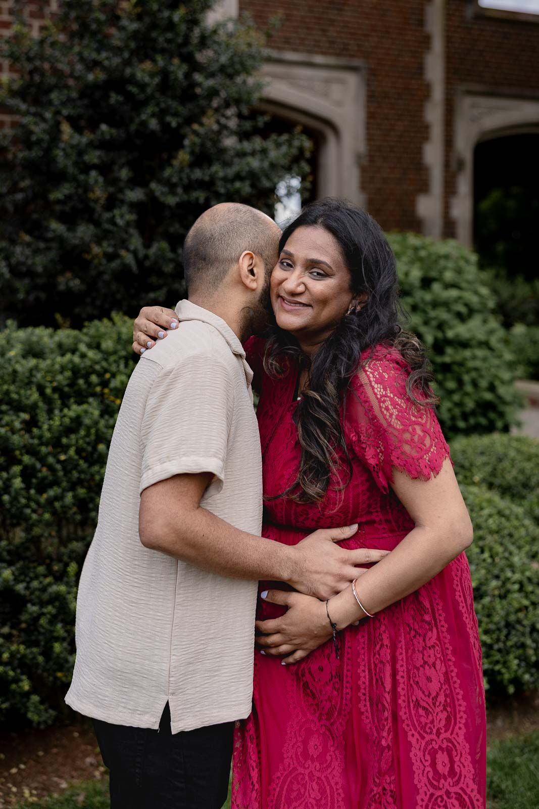 Intimate maternity moment between couple in red lace dress and beige shirt against brick building backdrop.