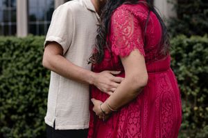 A couple embraces while standing in front of green hedges, with one person wearing a vibrant red lace dress.