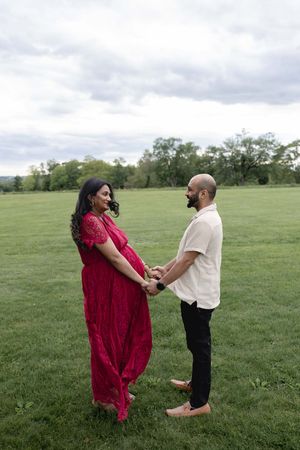A couple stands together in a green field with trees, the woman wearing a flowing red dress while holding hands.