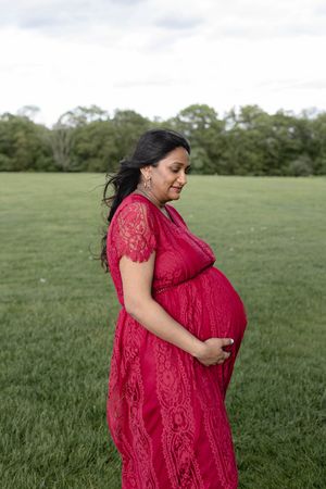 Expectant mother wearing red lace dress poses in green field during maternity photoshoot.