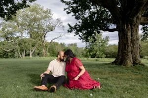 Two people in casual attire sit together on green grass under a large tree in a peaceful park setting.