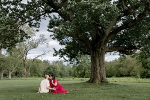 Two people in formal dresses sit together on grassy lawn beneath large tree on summer day.