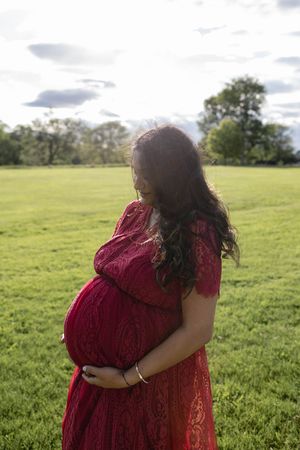 A pregnant person in a red lace dress stands in a sunlit meadow surrounded by green grass and trees.
