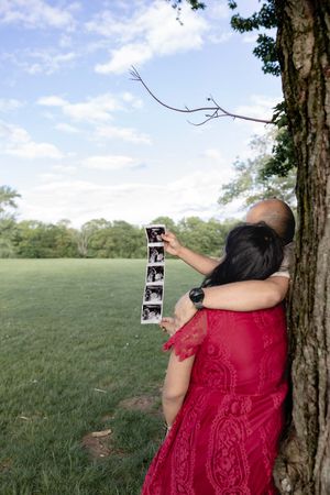 Someone in a red lace dress holds up photo strips while leaning against a tree in a grassy field on a sunny day.