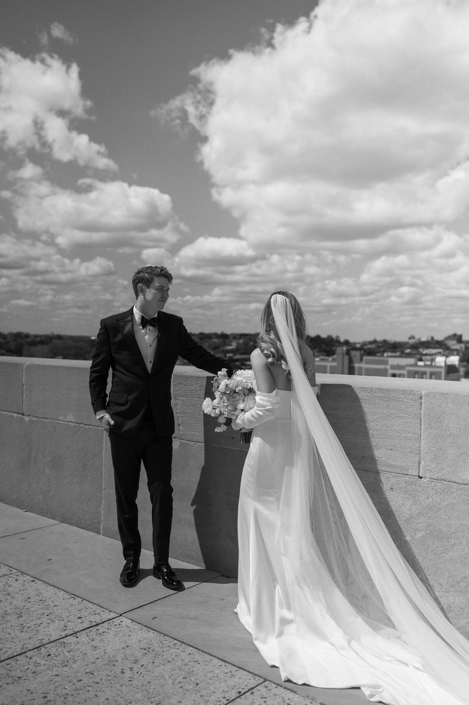 Black and white photo of newlyweds in formal attire standing on rooftop terrace with dramatic cloud-filled sky.