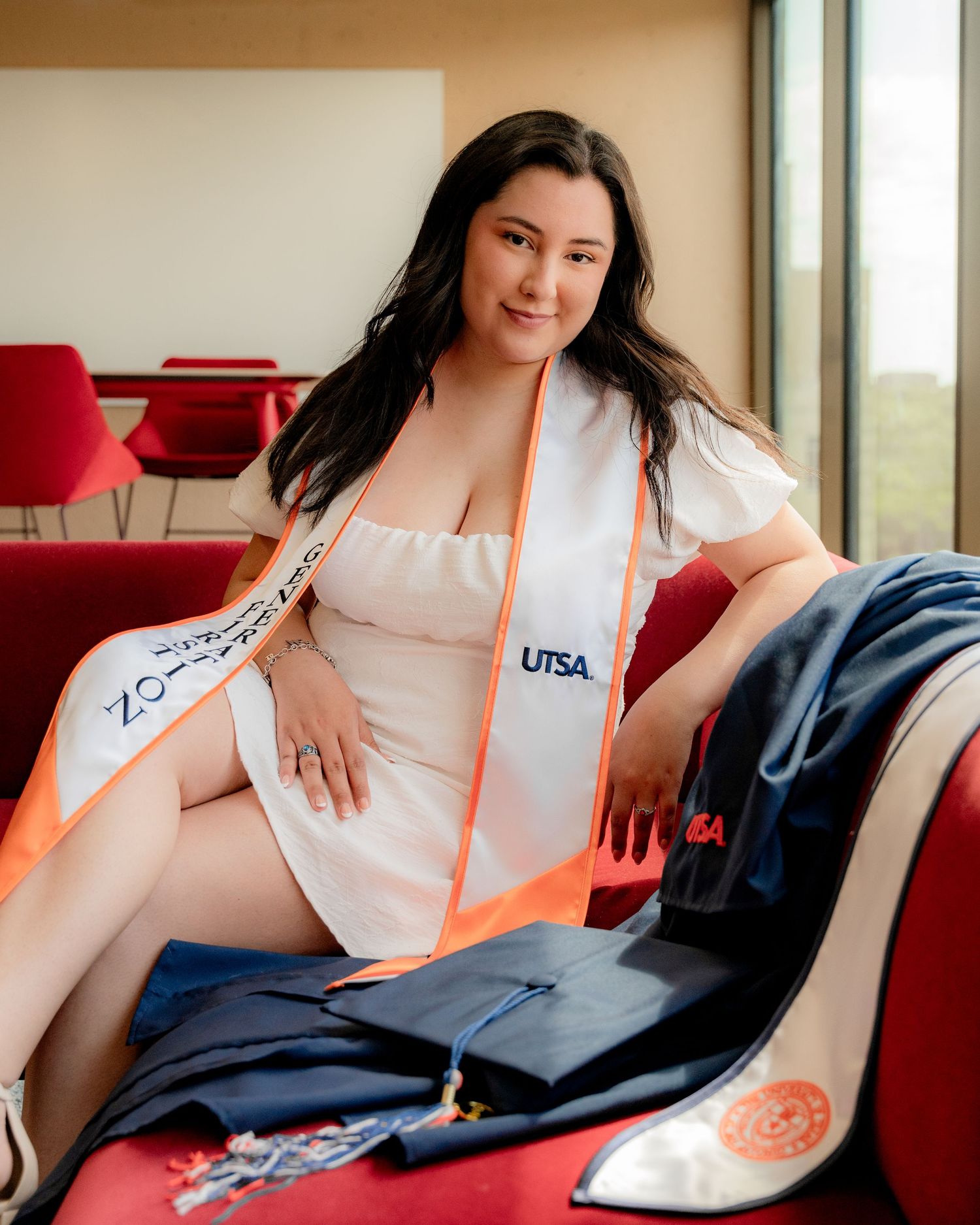 Graduate in white dress poses on red chair wearing UTSA graduation stole.