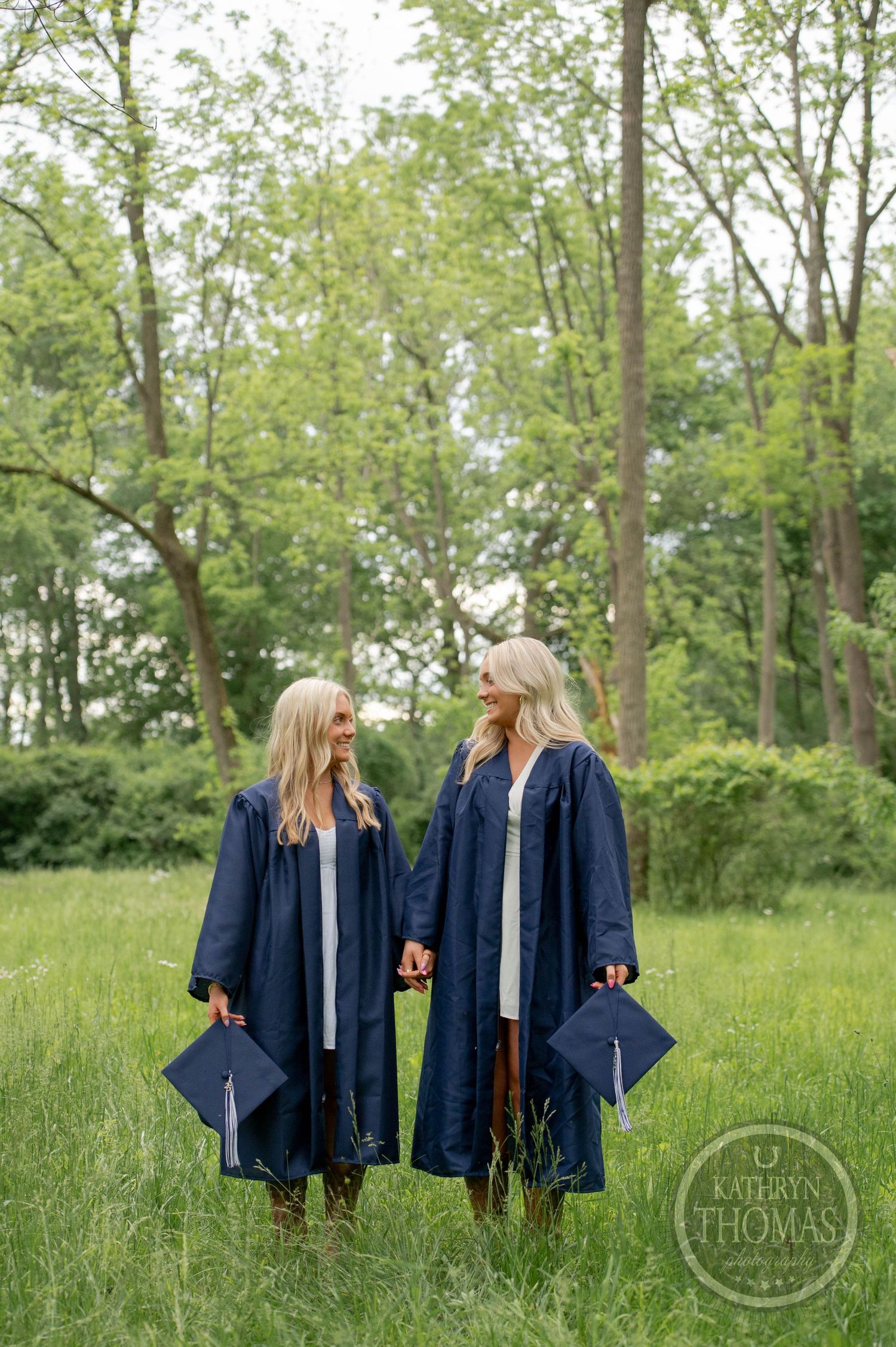 Two graduates in navy blue commencement gowns hold their caps while standing in a lush green field surrounded by trees.