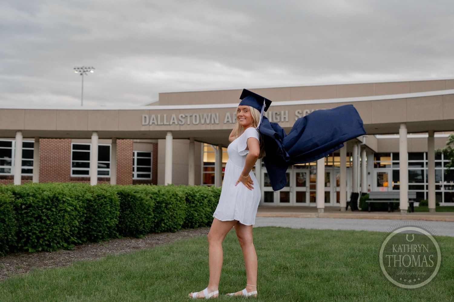 Graduate celebrates outside Bellwood High School entrance with dramatic dip pose in white dress and navy cap and gown.