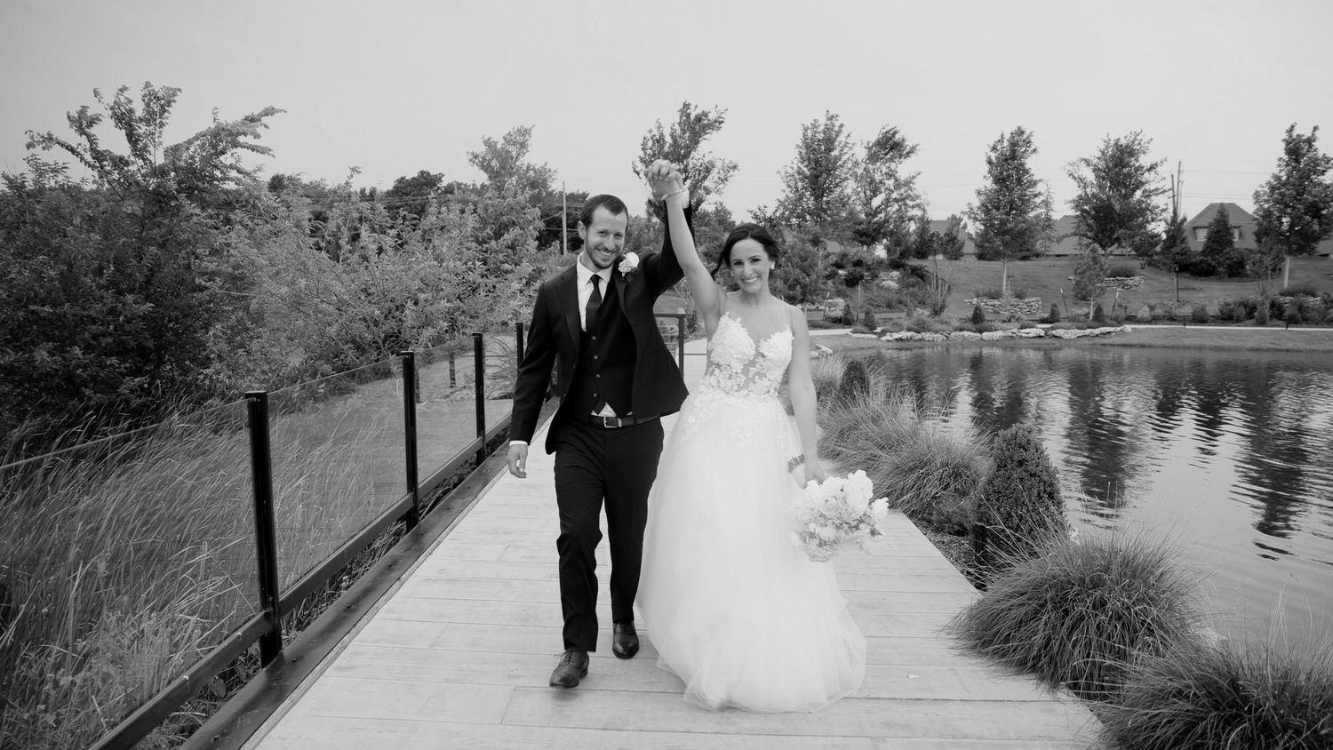 A happy couple walks along a peaceful lakeside path surrounded by lush greenery on their wedding day.