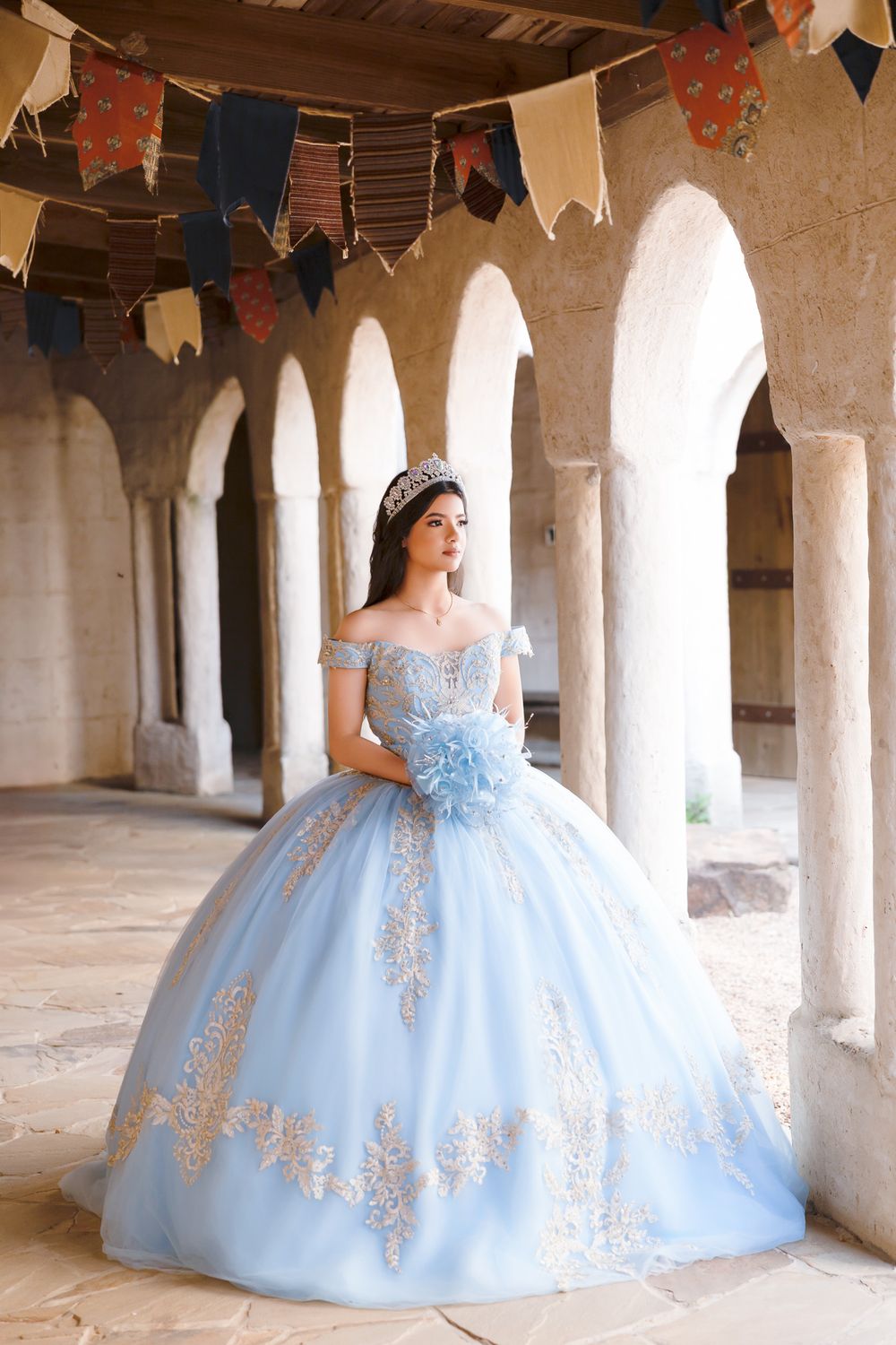 Light blue quinceañera ball gown with gold embroidery details photographed under stone archways with decorative bunting.