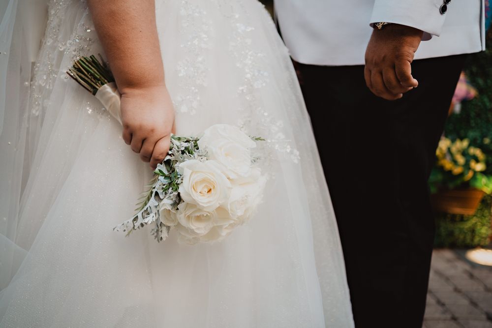 Close-up of a bride holding a delicate white rose bouquet next to a groom in black suit at an outdoor wedding ceremony.