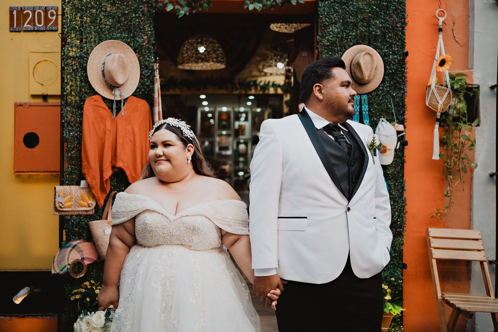 Couple in wedding attire holding hands against vibrant orange wall with hanging hats and greenery.