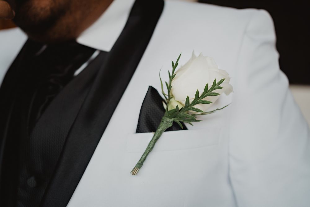 A white boutonniere with delicate greenery and black ribbon accent pinned to a crisp white suit lapel.