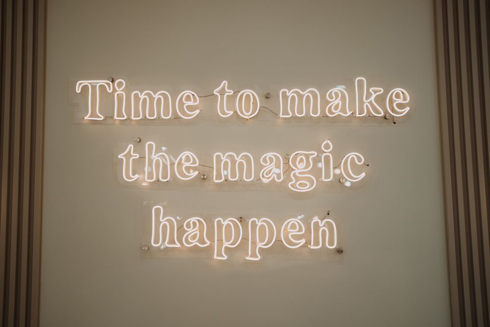 Glowing neon sign on wall reads 'Time to make the magic happen' in elegant cursive lettering.