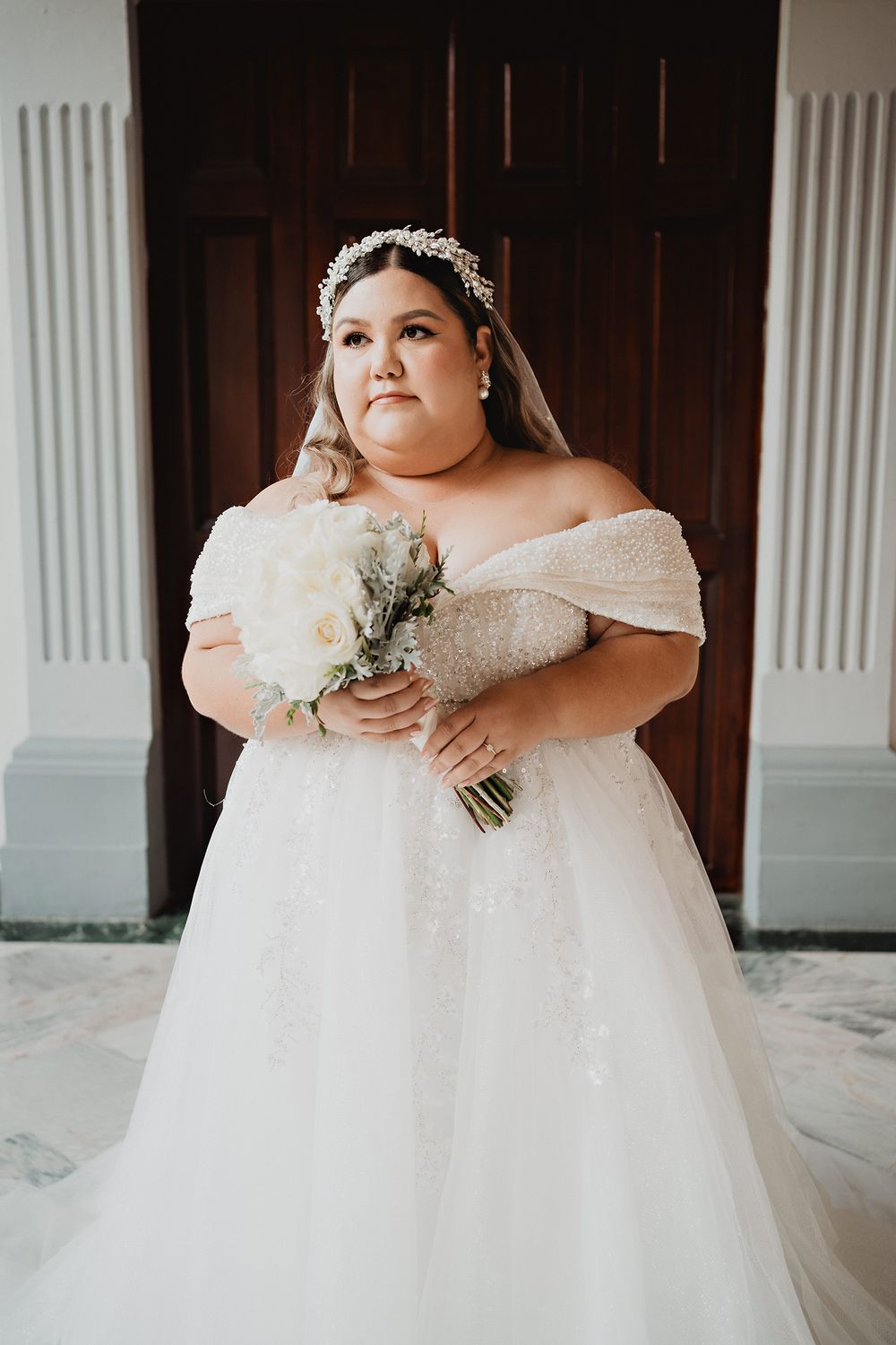 Bride in off-shoulder plus size ballgown holds white floral bouquet while posing against dark wooden door.